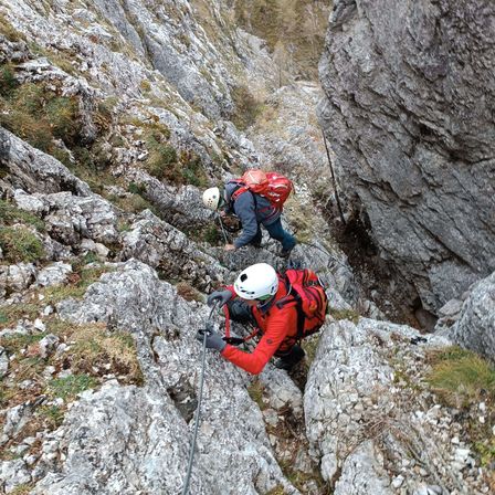 Zwei Kletterer erklimmen einen steilen felsigen Bergpfad und tragen Helme und Rucksäcke. Einer benutzt einen Kletterpickel und beide tragen Handschuhe.