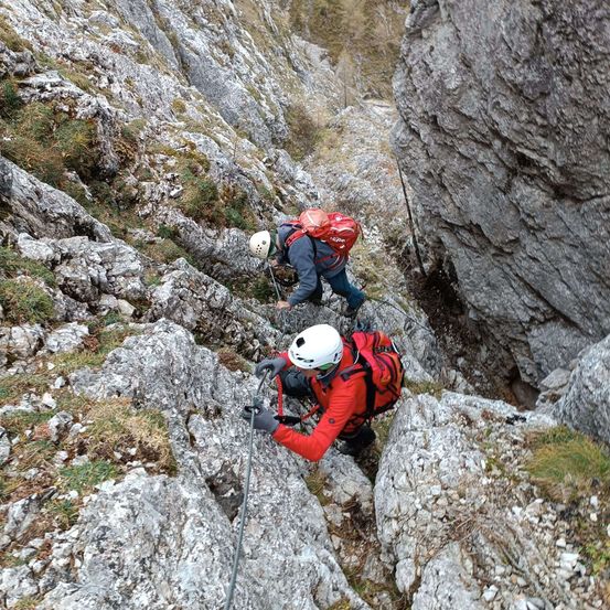 Zwei Kletterer erklimmen einen steilen felsigen Bergpfad und tragen Helme und Rucksäcke. Einer benutzt einen Kletterpickel und beide tragen Handschuhe.