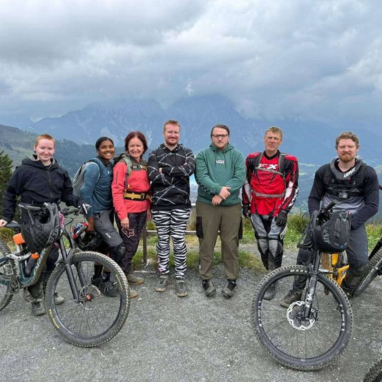 Eine Gruppe von Personen steht zusammen auf einem Bergpfad und posiert für ein Foto mit ihren Fahrrädern. Der Hintergrund besteht aus Bergen und bewölktem Himmel.