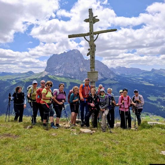 Eine Gruppe von Wanderern posiert für ein Foto auf einem Berggipfel mit einem großen hölzernen Kreuz. Die Gruppe steht auf saftigem Boden mit einer wunderschönen Bergkette im Hintergrund.