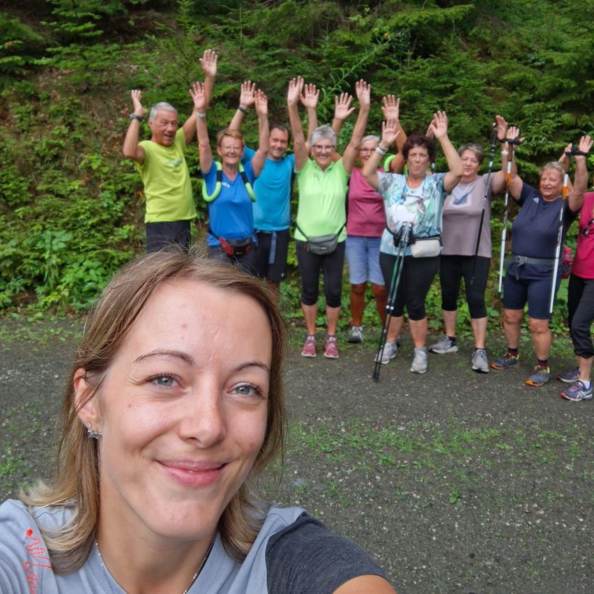 A group of hikers, all smiling and raising their hands, stand in a line on a gravel path with a forest backdrop.