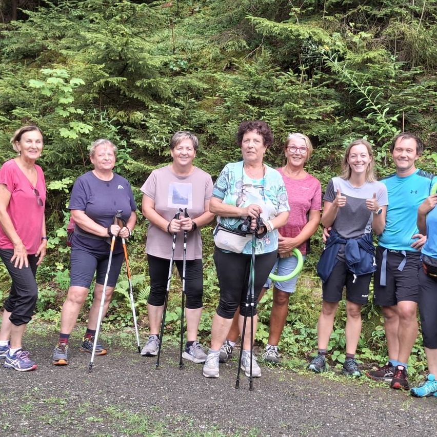A group of smiling individuals, dressed in casual outdoor attire, stand together on a gravel path with lush greenery in the background. They hold walking sticks and appear ready for a hike.