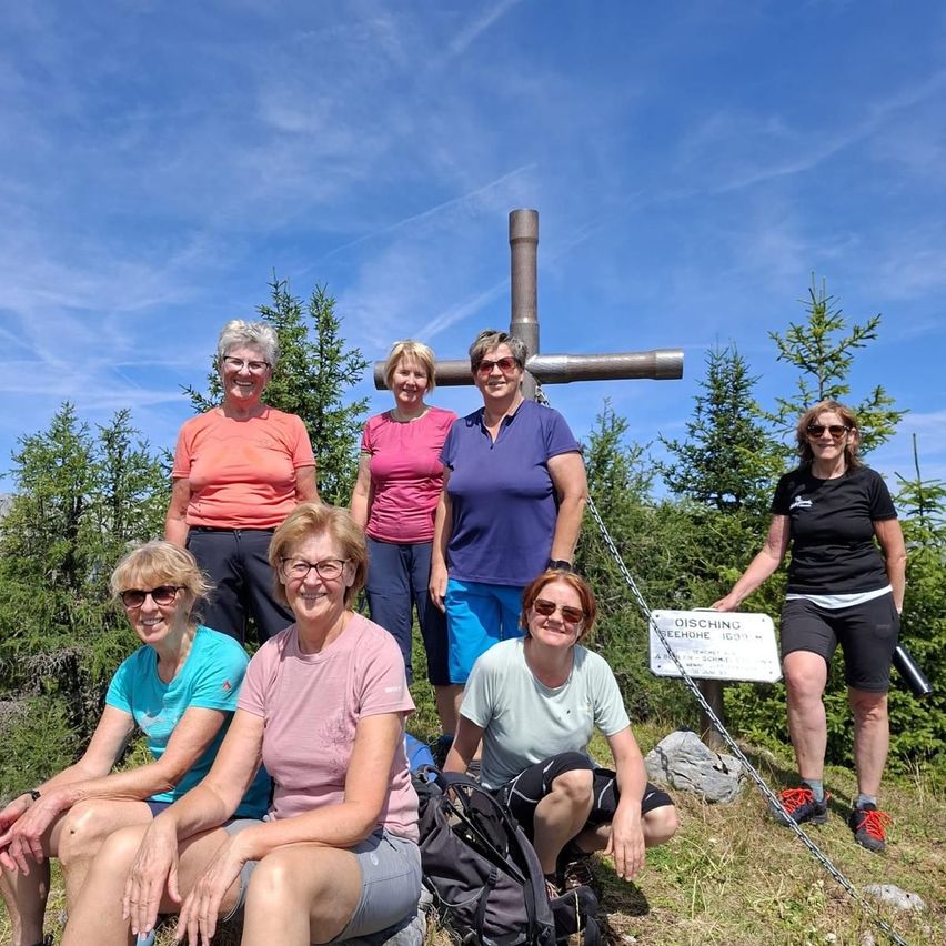 Eine Gruppe von Wanderinnen posiert für ein Foto auf einem Bergpfad mit einem Kreuz und einem Schild im Hintergrund.