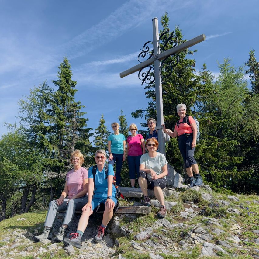 Eine Gruppe von Wanderern, in Freizeitkleidung gekleidet, posiert für ein Foto vor einem Kreuz auf einem Berg. Sie sind von Kiefern umgeben, unter einem blauen Himmel.