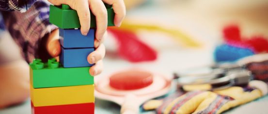 Eine Kinderhand stapelt bunte Lego-Steine. Der Tisch ist mit Spielzeug übersät. Im Hintergrund sind Handschuhe zu sehen.