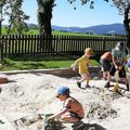 Eine Gruppe von Kindern spielt draußen in einem Sandkasten mit Schaufeln und einem Spielzeugauto. Bäume und ein Zaun sind im Hintergrund.