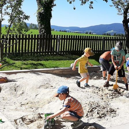 Eine Gruppe von Kindern spielt draußen in einem Sandkasten mit Schaufeln und einem Spielzeugauto. Bäume und ein Zaun sind im Hintergrund.