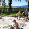 Eine Gruppe von Kindern spielt draußen in einem Sandkasten mit Schaufeln und einem Spielzeugauto. Bäume und ein Zaun sind im Hintergrund.