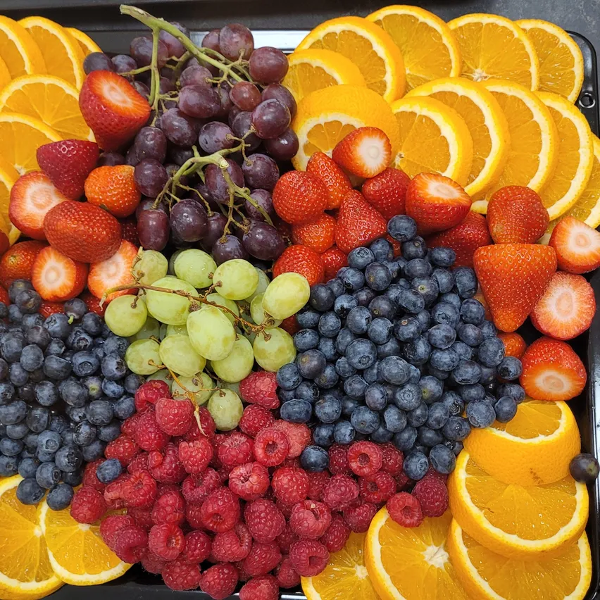 Ein Tablett mit verschiedenen Früchten, darunter Erdbeeren, Trauben, Blaubeeren, Himbeeren und Orangenscheiben. Die Früchte sind auf einem schwarzen Tablett farbenfroh arrangiert.
