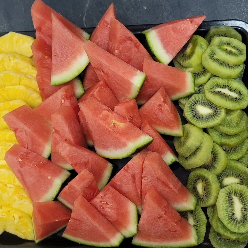 Ein schwarzer Teller mit verschiedenen geschnittenen Früchten, einschließlich Wassermelone, Kiwi und Ananas. Die Wassermelone ist rot mit Samen, Kiwi grün und Ananas gelb.