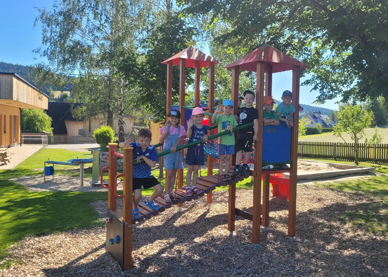 Eine Gruppe von Kindern spielt auf einer hölzernen Spielstruktur in einem Park, einige stehen auf einer Leiter, während andere auf einer Schaukel sitzen.