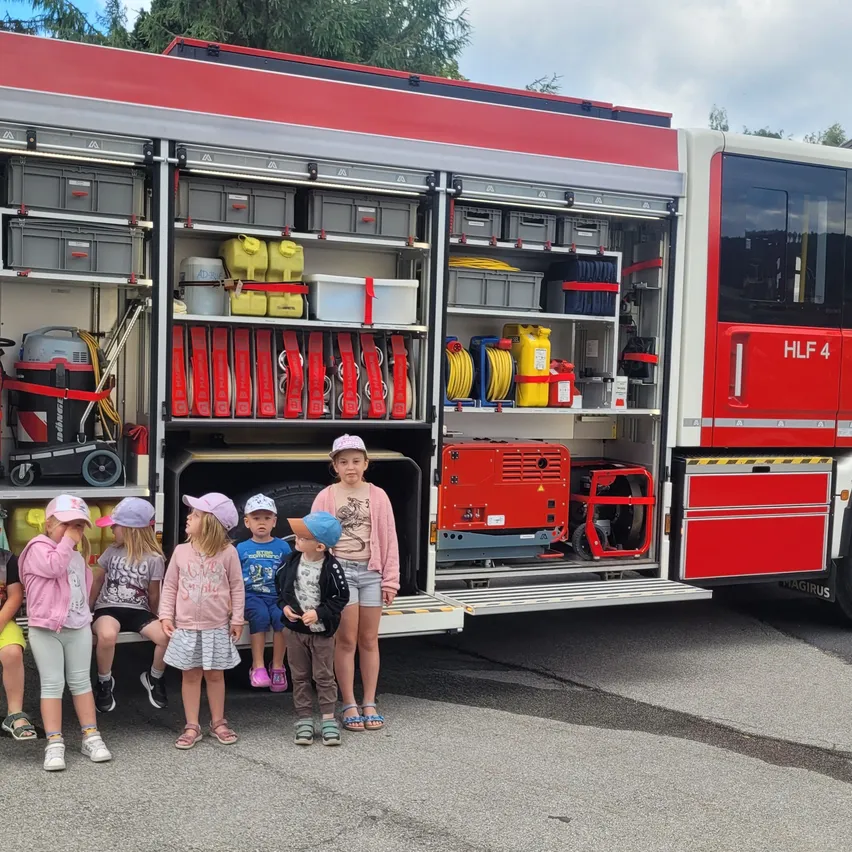 Eine Gruppe von Kindern steht vor einem Feuerwehrauto mit offenen Türen, das verschiedene Ausrüstungsgegenstände im Inneren zeigt. Die Kinder tragen Mützen und Turnschuhe.