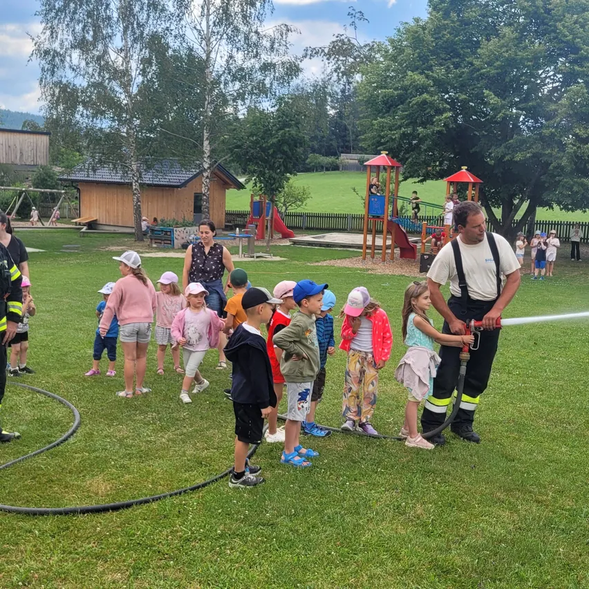 Eine Gruppe von Kindern versammelt sich auf einem Grasfeld mit einem Feuerwehrmann, der Wasser sprüht. Die Kinder stehen in einer Reihe mit Hüten und Turnschuhen. Dahinter befindet sich ein Spielplatz mit Rutschen und einem Zaun.