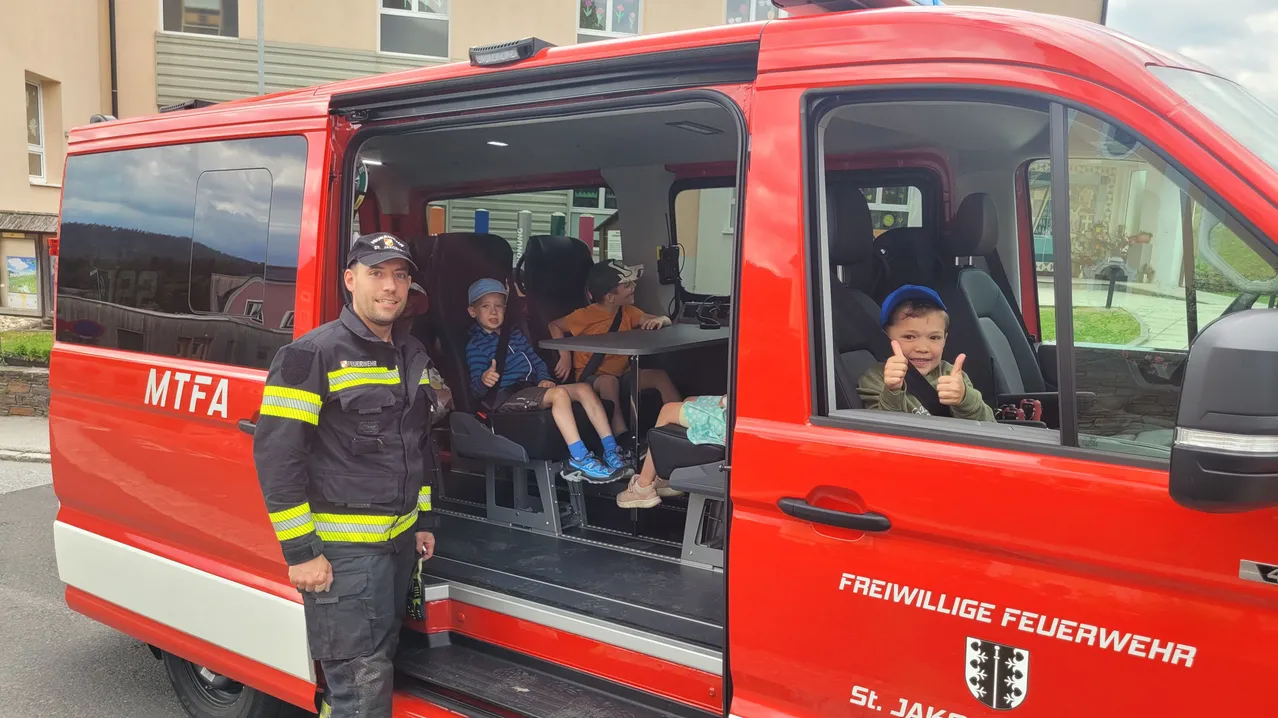 Ein Feuerwehrmann steht neben einem roten Feuerwehrauto, in dem drei Kinder sitzen, alle mit Sicherheitsgurten angeschnallt und lächelnd. Das Auto trägt die Aufschrift 'Freiwillige St. Jakob.'