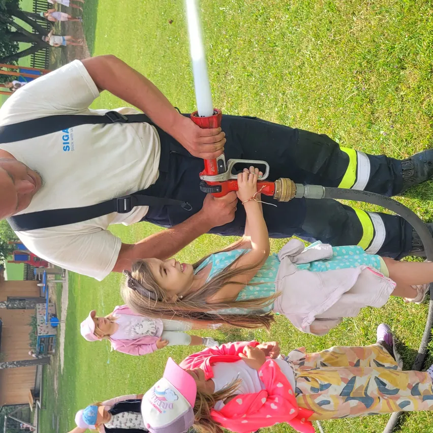 Ein Feuerwehrmann demonstriert einer Gruppe von Kindern an einem sonnigen Tag die Benutzung einer Feuerwehrspritze.