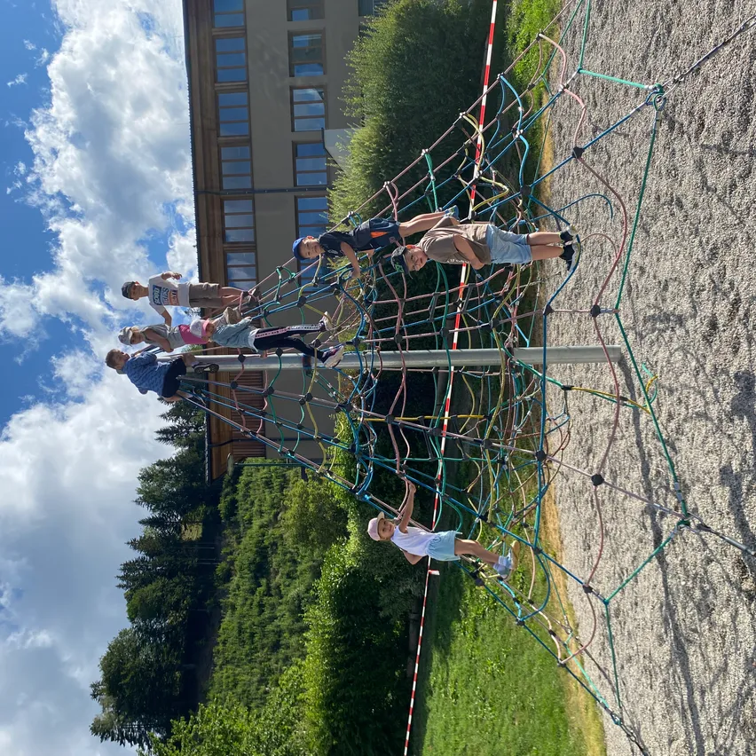 Mehrere Kinder klettern eine bunte Seilleiter auf einem Spielplatz. Sie sind draußen in einer sonnigen Umgebung, mit einem Gebäude und Bäumen im Hintergrund.