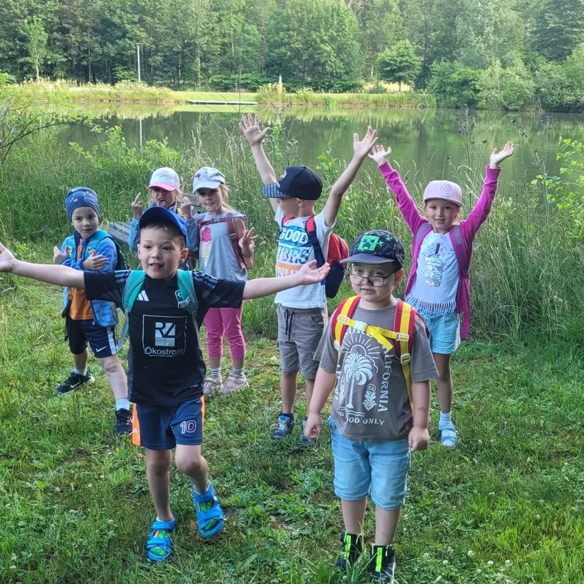 Eine Gruppe von Kindern steht in einem Grasfeld in der Nähe eines Teichs, mit Bäumen und Grünpflanzen im Hintergrund. Sie tragen Hüte, Rucksäcke und Turnschuhe, einige mit erhobenen Armen.