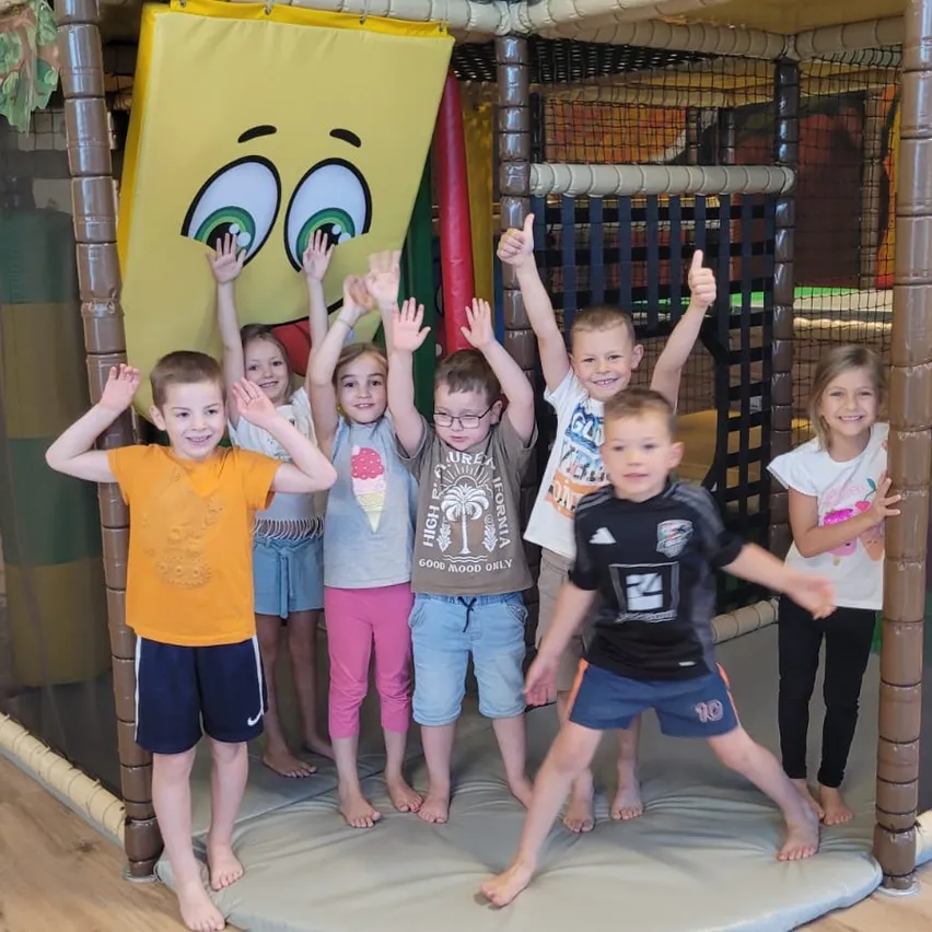 Eine Gruppe von Kindern in einem Spielbereich, posiert mit erhobenen Händen. Sie stehen in der Nähe eines gelben Aufblasbaren, hinter ihnen ist eine Netzwand zu sehen.