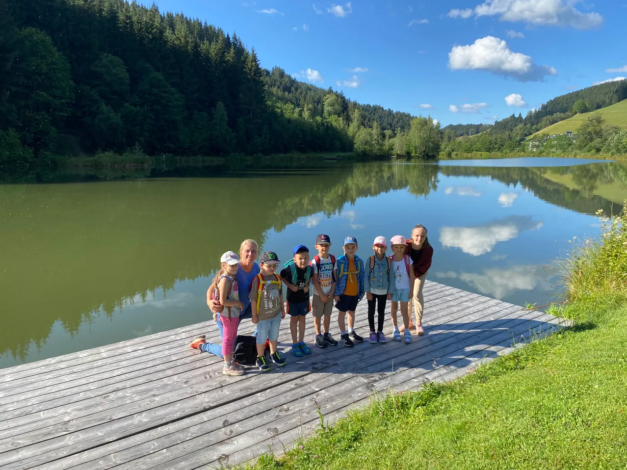 Eine Gruppe von Kindern und Erwachsenen posiert auf einem Steg am See mit einer Kulisse aus Hügeln und blauem Himmel.