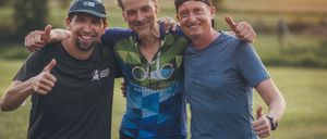 Drei Männer stehen gemeinsam auf einem Feld, lächeln und geben Daumen hoch. Der Mann in der Mitte trägt ein Radtrikot mit Logo.