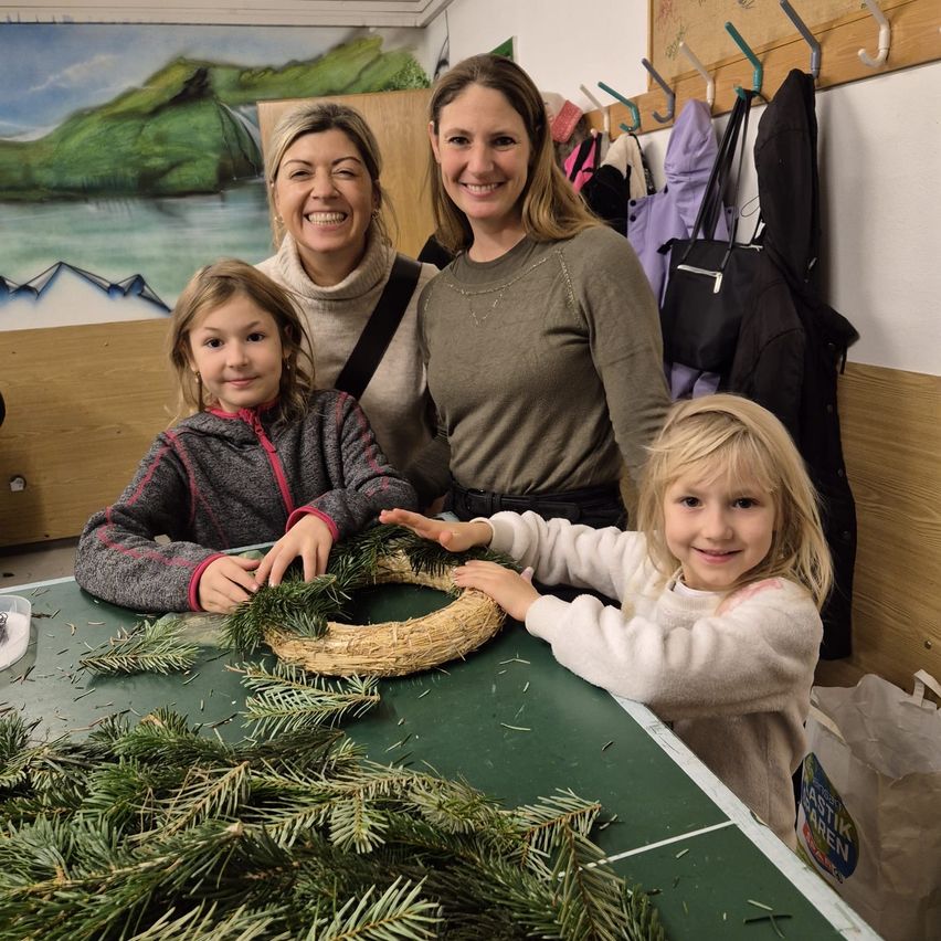 Drei Frauen und zwei Mädchen sind in einem Raum, lächeln und posieren für ein Foto. Die Mädchen halten einen Kranz, und die Frauen stehen hinter ihnen. Es gibt eine Wand mit einem Gemälde, einen Tisch mit einem Kranz und einige hängende Kleidungsstücke.