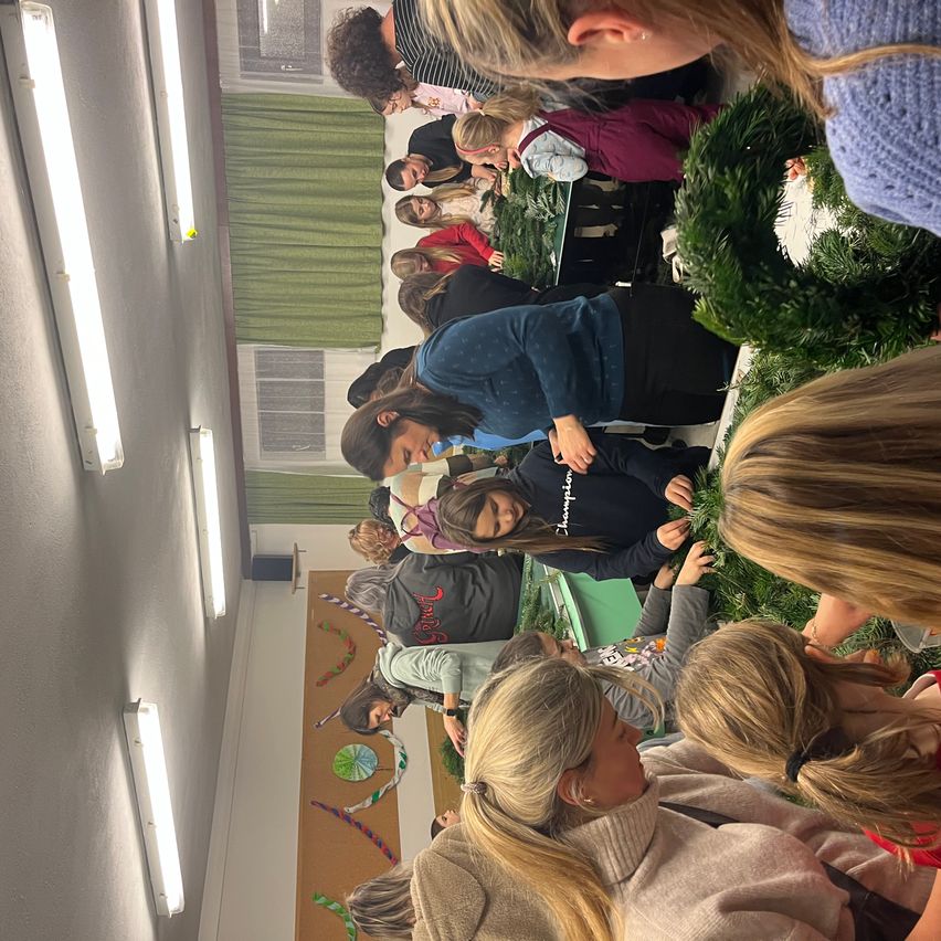 Eine Gruppe von Menschen versammelt sich in einem Raum und arbeitet an Kränzen. Der Raum hat weiße Wände, grüne Vorhänge und verschiedene Dekorationen. Sie stehen und beugen sich über Tische, konzentriert auf ihr Handwerk.