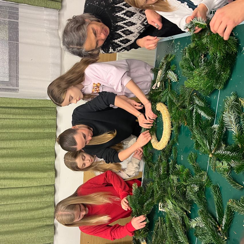 Eine Gruppe von Kindern und einem Erwachsenen schmücken einen grünen Kranz mit künstlichen Tannenzweigen auf einem Tisch.