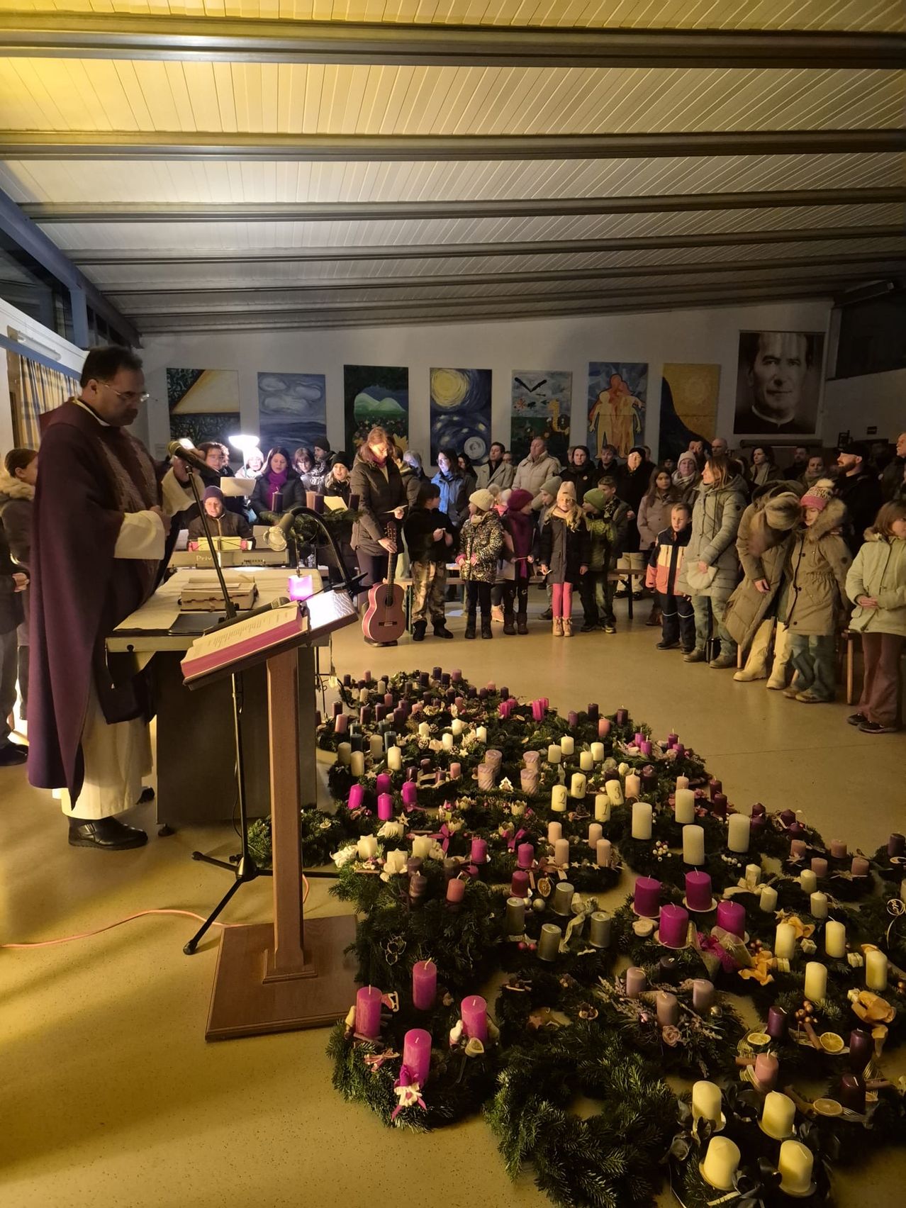 Ein Priester leitet eine Zeremonie in einem Saal mit vielen versammelten Menschen. Er steht an einem Pult mit einem Buch und einem Mikrofon. Der Boden ist mit zahlreichen Kerzen und Kränzen geschmückt.
