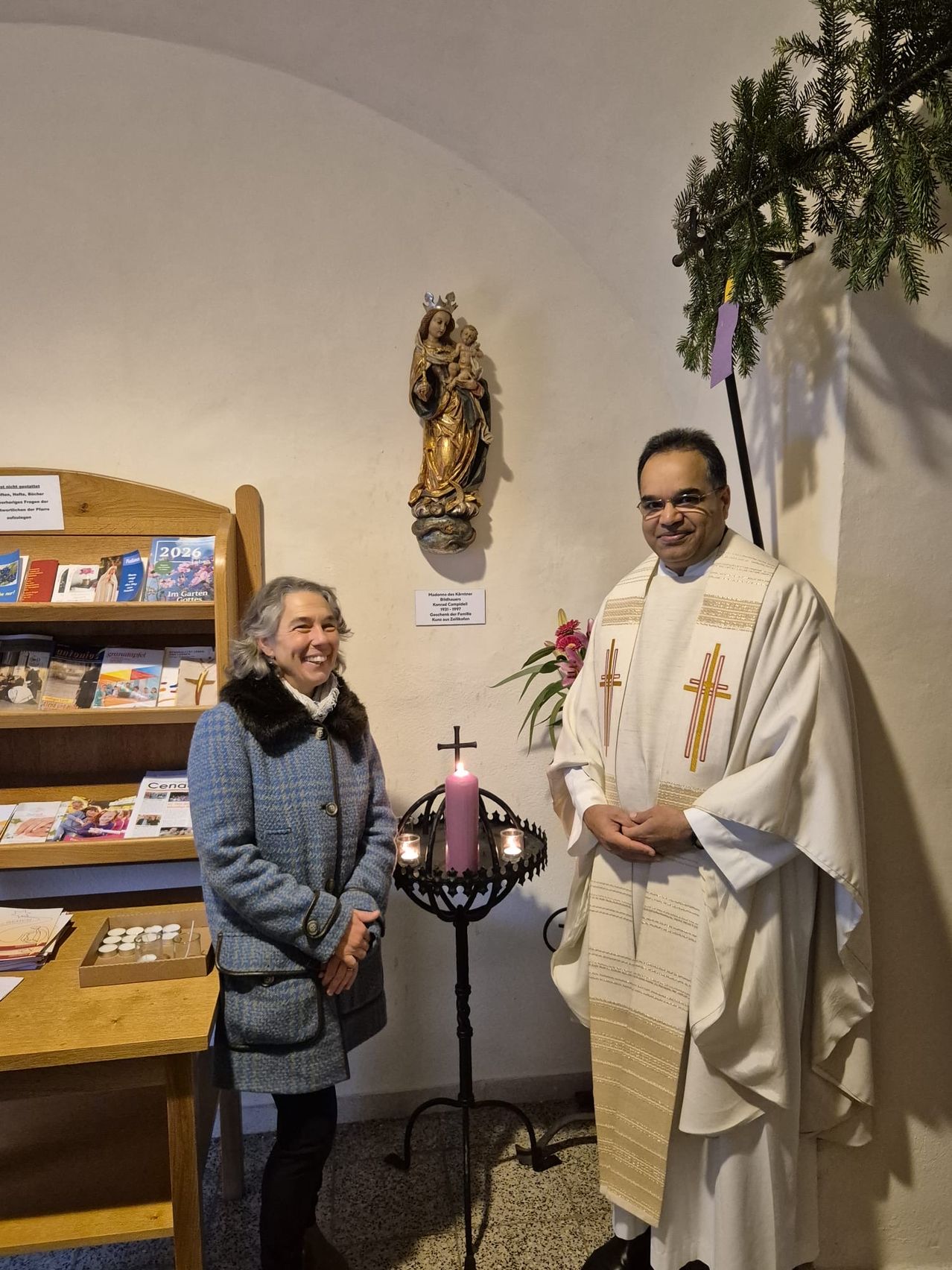 Eine lächelnde Frau in einem Mantel steht neben einem Priester in einer weißen Robe, beide posieren für ein Foto. Hinter ihnen steht ein Kerzenhalter, ein Bücherregal und eine Statue einer Heiligen.