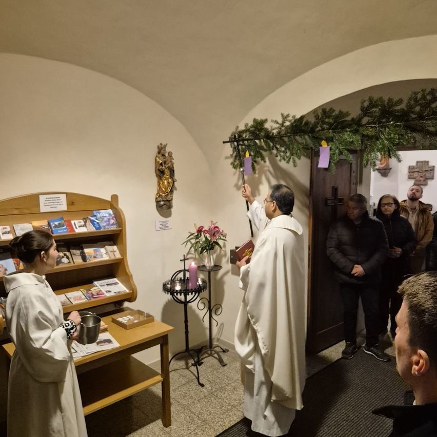 Ein Priester hält eine Kerze in einer Kapelle, eine Frau hält eine Schüssel. Ein Regal mit Büchern, ein Kerzenständer und eine Pflanze im Hintergrund. Mehrere Menschen beobachten.