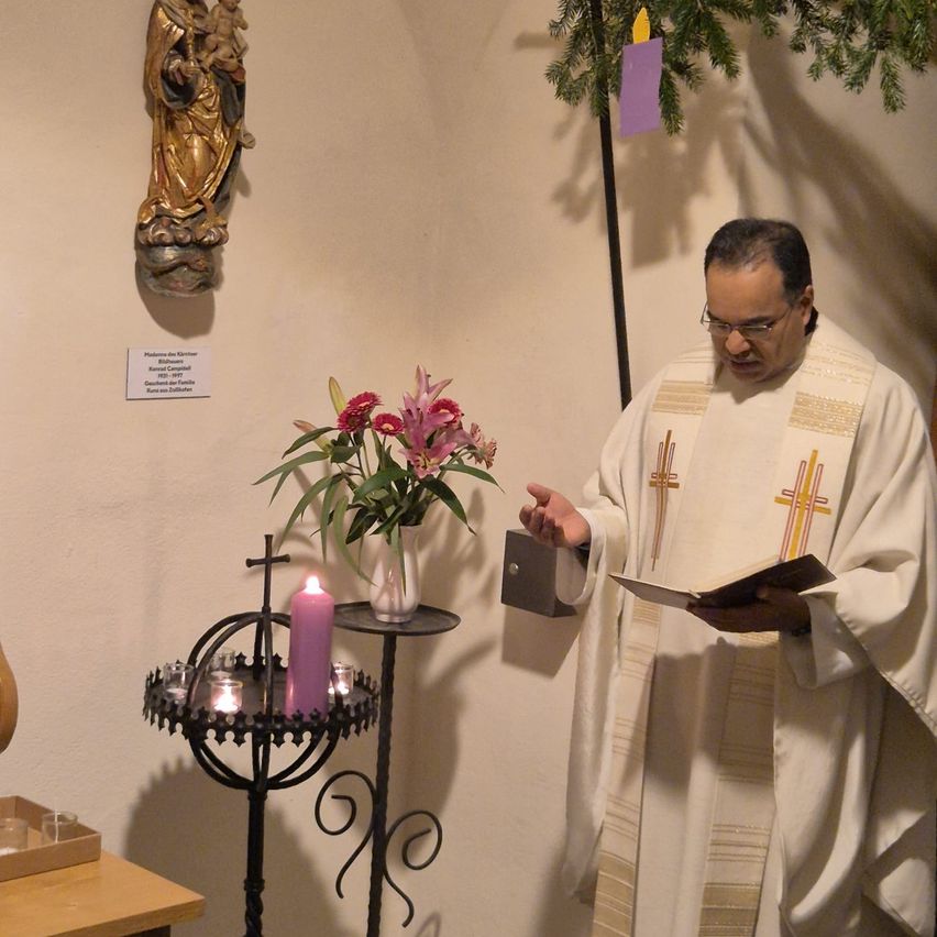 Ein Priester steht in einem Raum und liest aus einem Buch. In der Nähe befindet sich ein Kerzenständer mit Blumen, einem Kreuz und einer Statue der Maria.