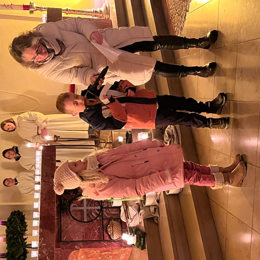 Eine Gruppe von Menschen ist in einer Kirche, zwei Jungen stehen und halten Mikrofone, während eine Frau hinter ihnen steht. Der Junge auf der rechten Seite trägt einen schwarzen Mantel, und ein Mädchen steht vor ihnen.