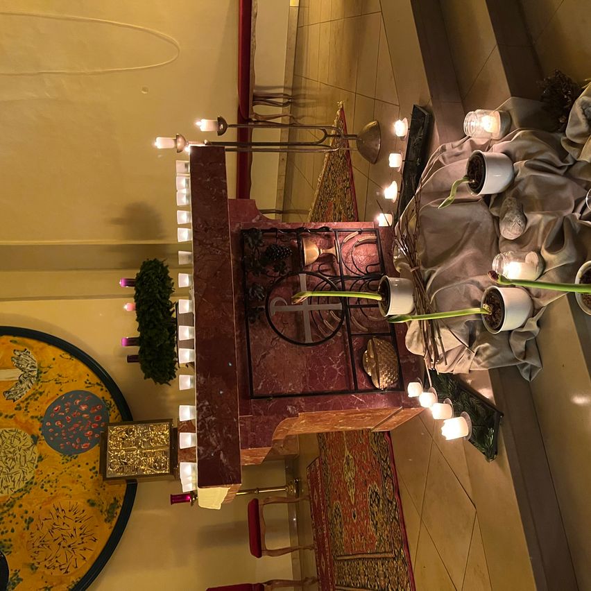 Auf einem Marmoraltar sind verschiedene Kerzen und Topfpflanzen angeordnet. Links befindet sich ein kreisförmiges Kunstwerk mit verschiedenen Designs. Der Boden ist gefliest und in der Nähe stehen rote Stühle.