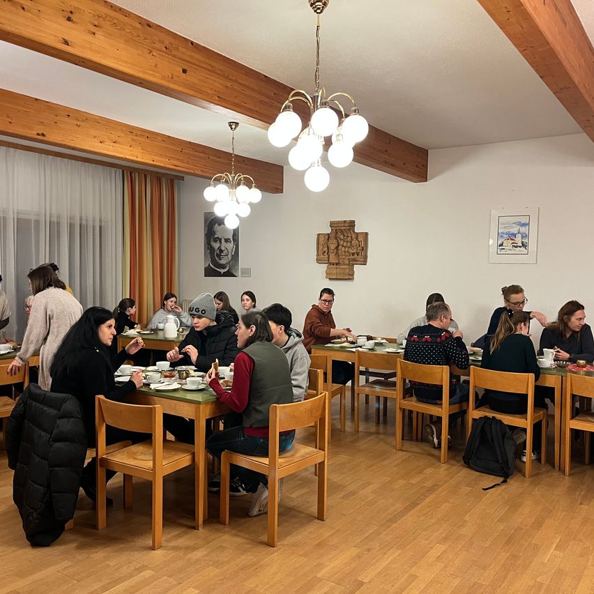 Eine Gruppe von Menschen sitzt an Tischen in einem Speisesaal. Der Raum hat Holzböden und -wände, mit Bilderrahmen und einer hängenden Kronleuchter.