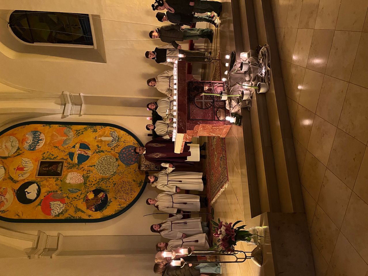 Auf einer Bühne stehen mehrere Menschen in weißen Gewändern vor einem Tisch, der mit Kerzen und Blumen geschmückt ist. Die Szene spielt sich in einer Kirche ab, an deren Wand ein farbenfrohes Wandbild zu sehen ist.