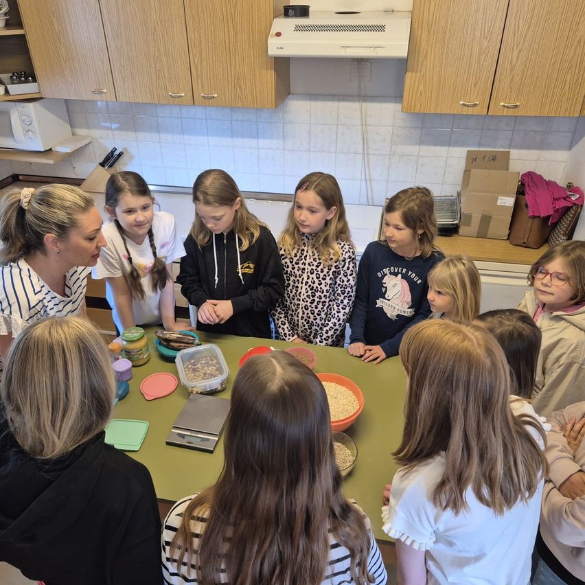 Eine Gruppe von Mädchen und einer Frau in einer Küche, die sich um einen Tisch mit verschiedenen Lebensmitteln und einer Waage versammelt haben.