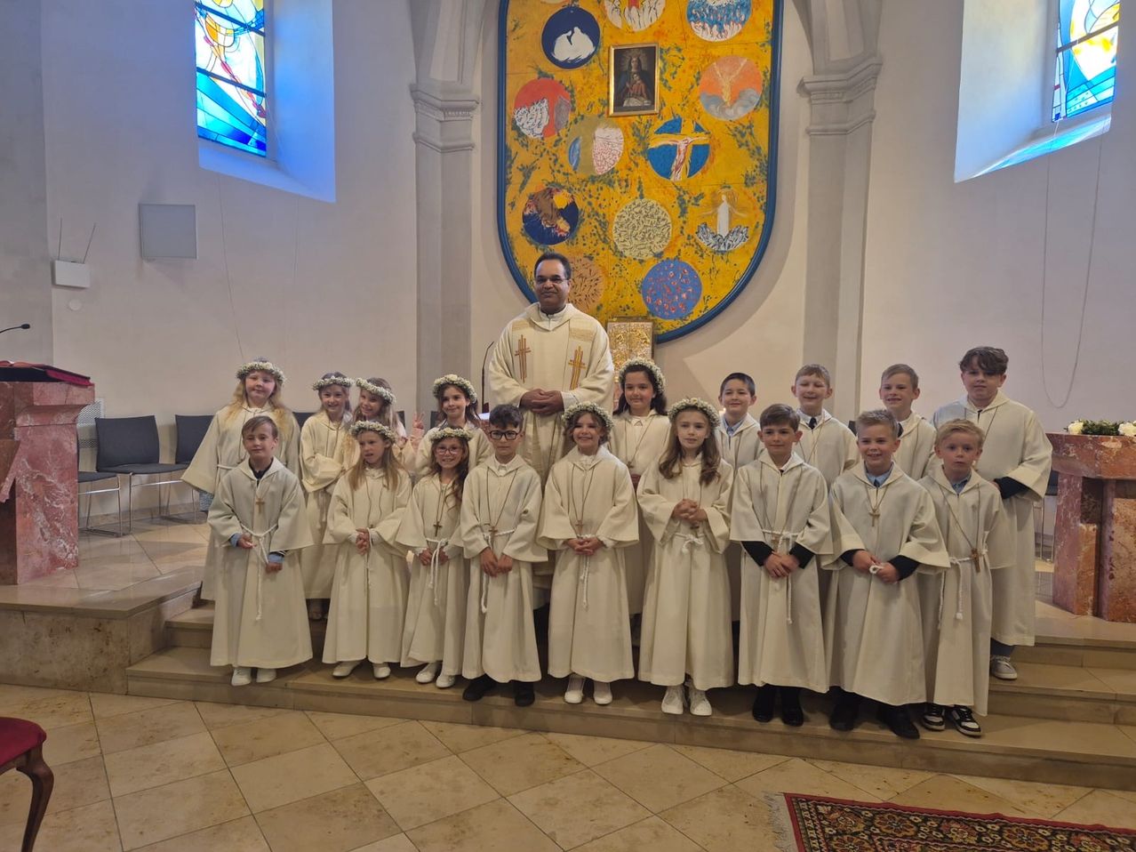 Ein Priester steht vor einem Kirchenaltar mit einer Gruppe von Kindern in weißen Gewändern. Die Kinder haben Blumen auf dem Kopf. Hinter dem Priester ist eine große Wandmalerei mit einem Bilderrahmen.