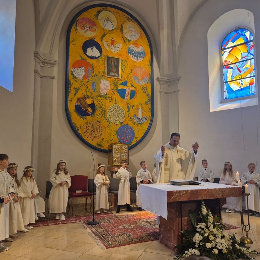 Ein Priester führt eine Zeremonie am Altar einer Kirche durch. Kinder in weißen Gewändern stehen in der Nähe und eine farbenfrohe Wandmalerei schmückt die Wand.