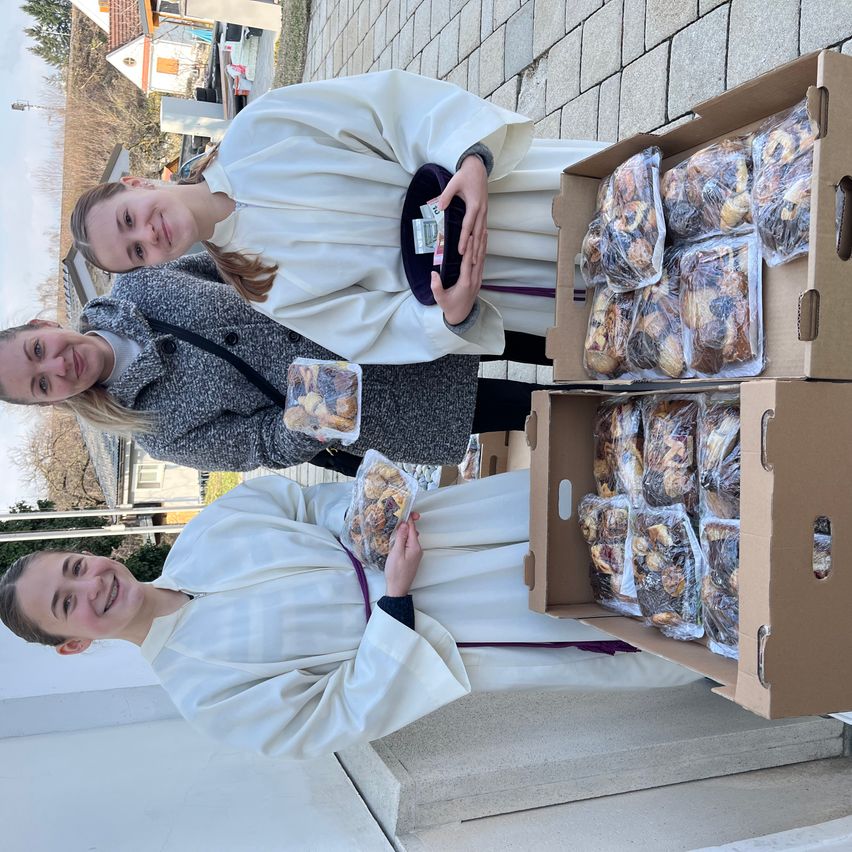 Zwei Frauen in religiöser Kleidung posieren mit Lebensmittelpaketen in Plastikbehältern und Pappkartons. Eine hält ein kleines Paket und die andere ein größeres. Sie lächeln und stehen neben den Kartons.
