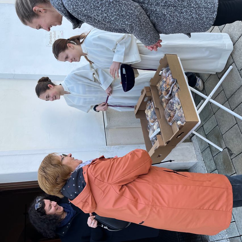 Mehrere Frauen in weißen Roben stehen neben einer Box mit Snacks, während andere zuschauen. Der Ort scheint eine Straßenecke zu sein.