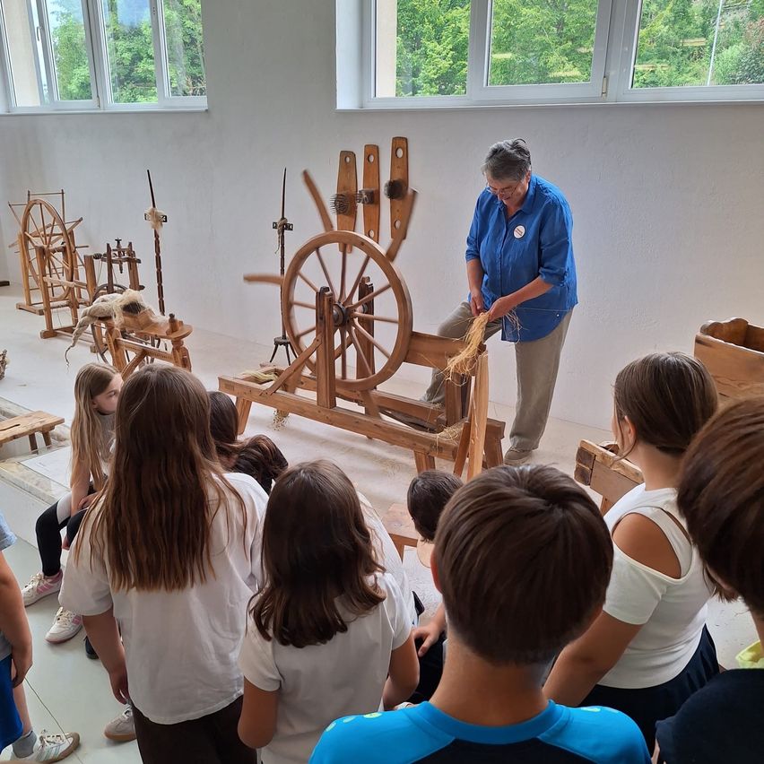 An elderly woman is demonstrating the use of a wooden spinning wheel to a group of children in a room with white walls and large windows.