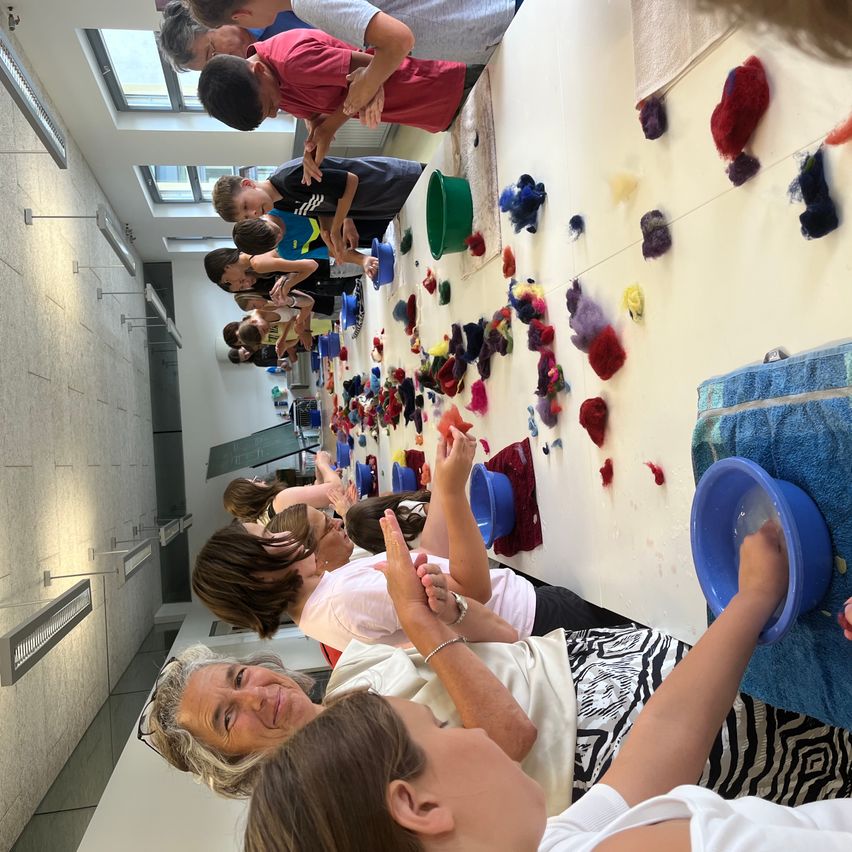 A group of people, including children and adults, are engaged in an activity involving colorful wool in a room with a white wall and a ceiling with skylights.