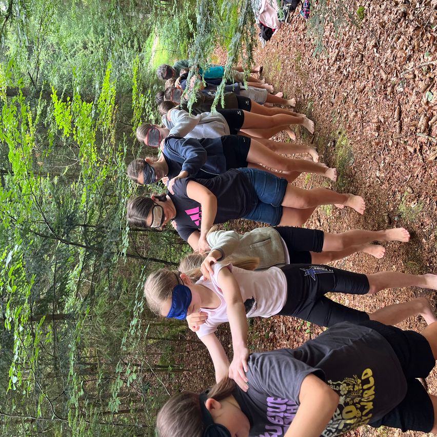 A group of people are standing in a forest, wearing blindfolds, and forming a line. They are likely participating in a team-building activity.
