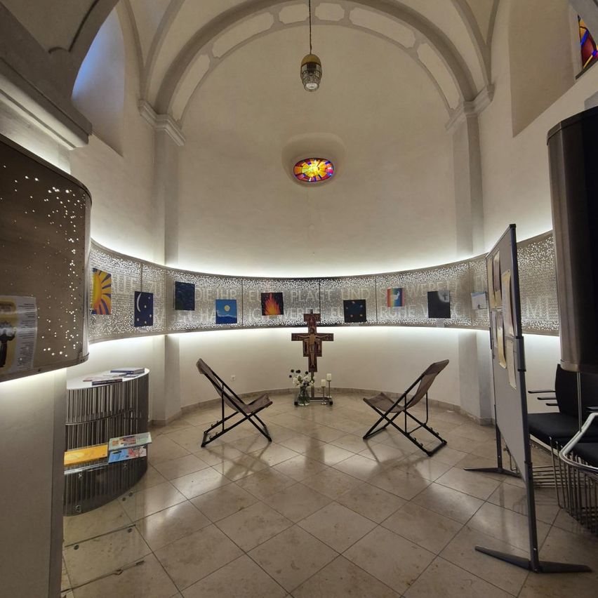 A small chapel with a cross at the center, surrounded by chairs and a shelf with books. The walls are adorned with various artworks.