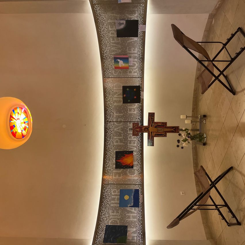 An aerial view of a chapel with a cross, candles, and artwork on the wall. Two folding chairs are placed on the right side.