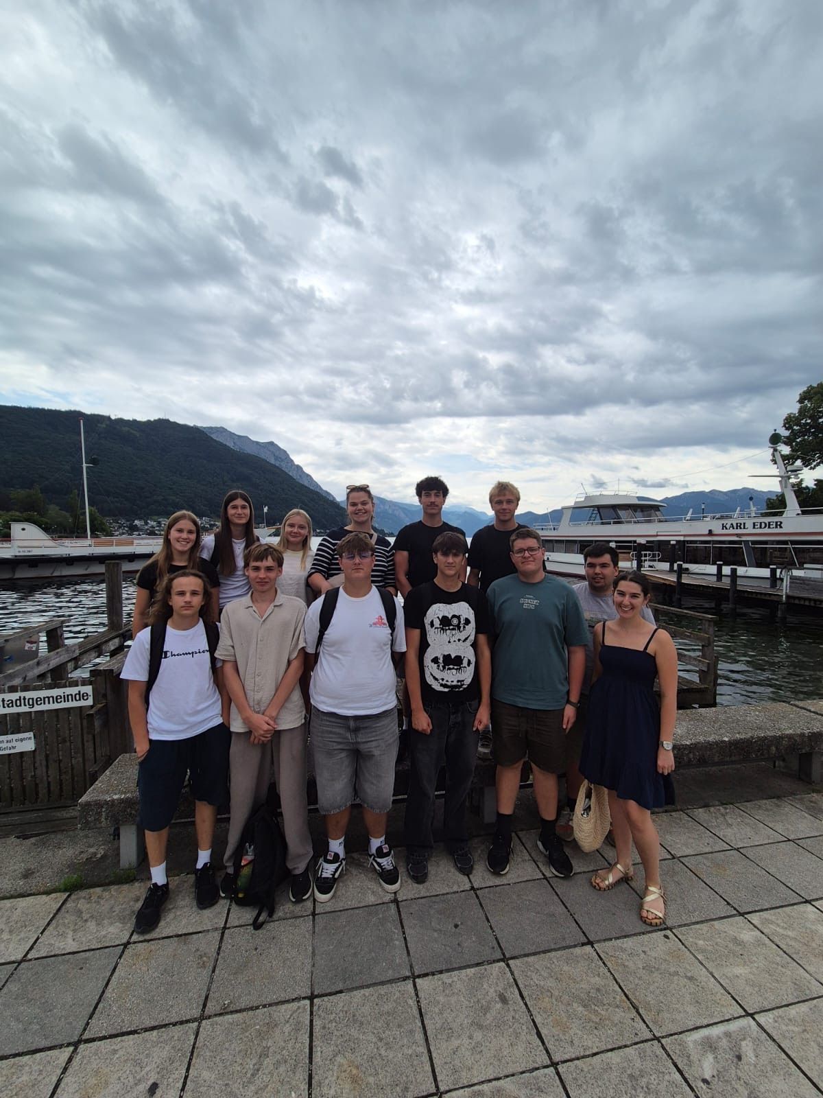 A group of young people stands on a pier by the water, with mountains and a boat in the background.