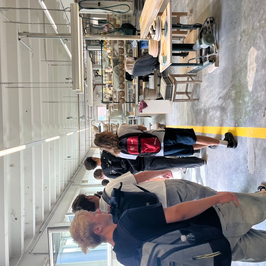 A group of people with backpacks are walking through a workshop with yellow lines on the floor.