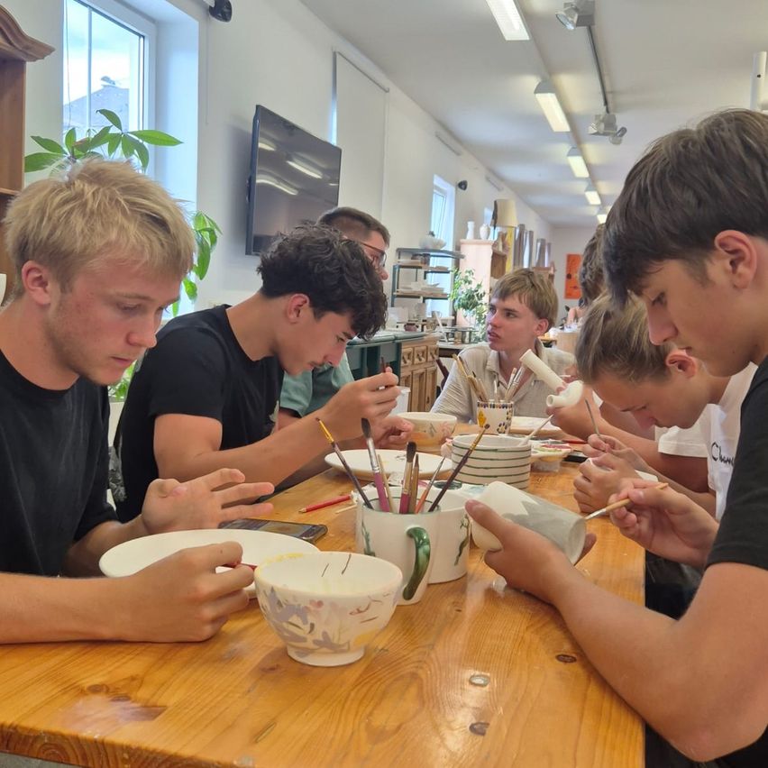 A group of young men are seated around a table, engaged in painting ceramic mugs. Each has a mug in front of them with brushes and paint. The room has a warm, natural light.