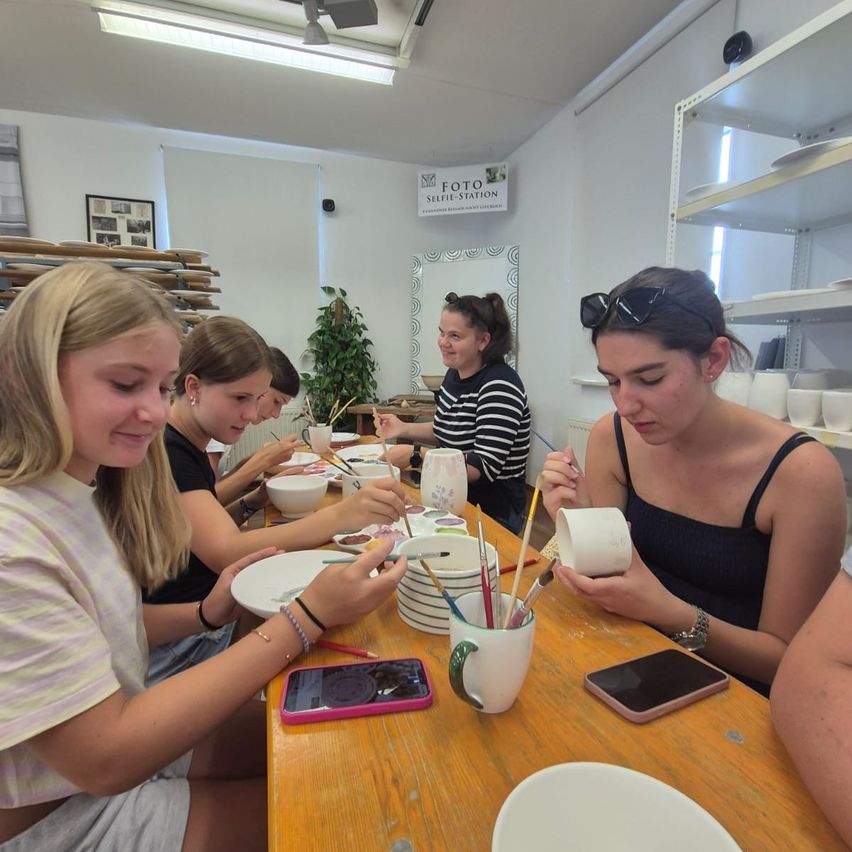 A group of women are seated at a table, engaged in a pottery painting activity. Each has a cup and paintbrush, with bowls and plates on the table. A sign in the background reads 'FOTO SELFIE-STATION'.