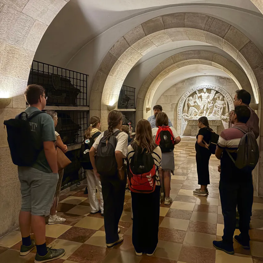 Eine Gruppe von Menschen, einige mit Rucksäcken, stehen in einem historischen Tunnel mit gewölbten Decken und gemustertem Boden und betrachten eine Steinwand mit einer alten Schnitzerei.